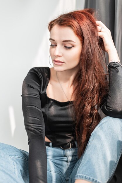 Modelo de chica bastante glamorosa en camiseta negra y jeans azules se sienta en el estudio con luz solar
