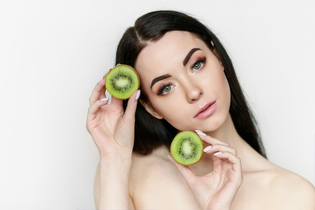 modelo com maquiagem natural segura dois pedaços de kiwi perto do rosto