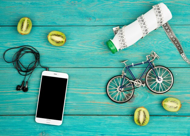 Modelo de bicicleta fones de ouvido smartphone kiwi garrafa de água e fita de centímetro na mesa de madeira azul
