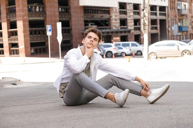 Modelo de cara com um corte de cabelo elegante, posando ao ar livre em uma camisa branca e calça cinza. penteado moderno fica perto de um moderno centro de negócios. Cara atraente na rua num dia de verão.