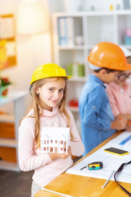 Foto modelo de casa. linda garota com um capacete segurando uma pequena maquete de casa em pé perto de colegas de classe