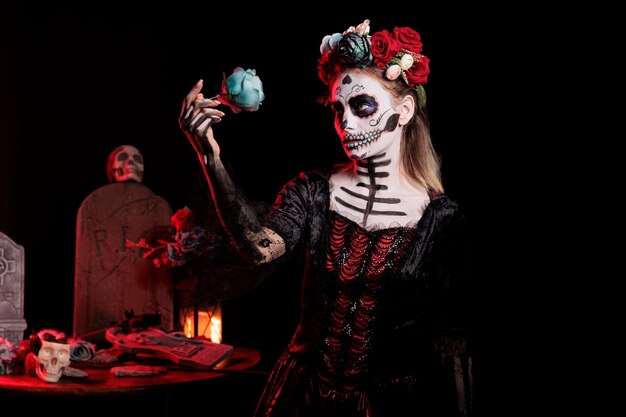 Modelo de glamour usando maquiagem preto e branco e coroa de flores para celebrar o ritual mexicano de halloween. Deusa da morte com fantasia de carnaval de horror e arte corporal, segurando a rosa.