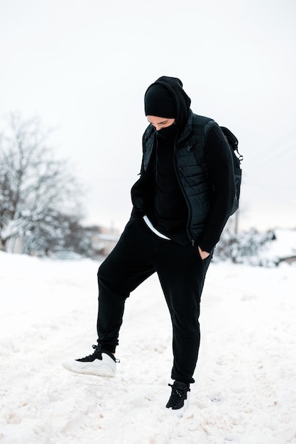 Modelo de hipster de cara elegante em agasalhos elegantes pretos e tênis brancos com uma mochila posa na neve em um dia frio de inverno.