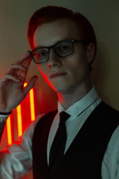 Modelo de jovem bonito e elegante com roupas elegantes de negócios com camisa e gravata endireita os óculos com luz de néon vermelha