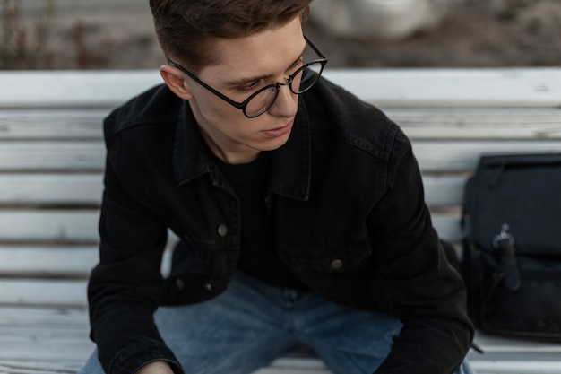 Modelo de jovem retrato elegante com penteado na moda na jaqueta jeans preta da moda em óculos vintage com mochila de couro na rua da cidade. um cara atraente está sentado no banco de madeira.