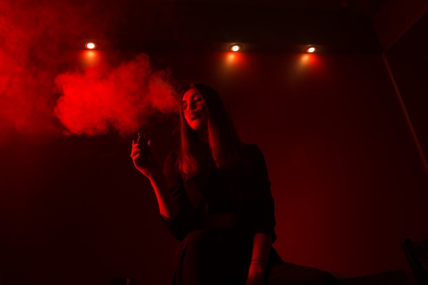 Modelo de menina fuma vape em luz vermelha.