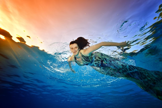 Modelo de menina nada e posando debaixo d'água na piscina em um vestido no contexto de um pôr do sol