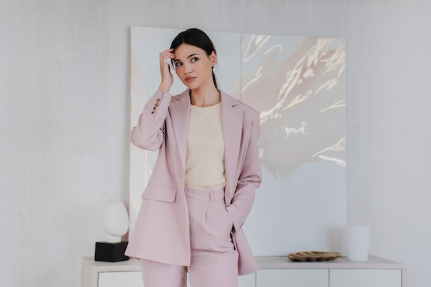 Modelo de moda bonita posando de tocar o cabelo vestido com terno rosa claro em pé na sala de estar Atraente jovem adulta asiática olhando de lado Linda elegante garota coreana se preparando para entrevista