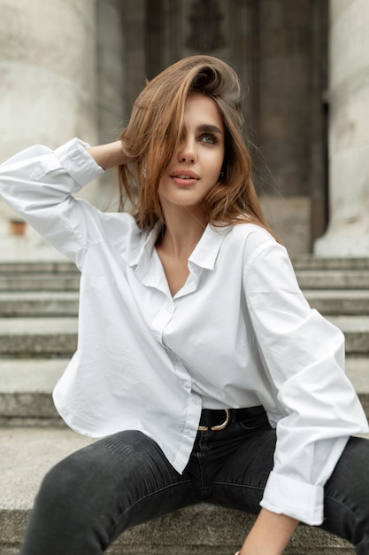 Modelo de moda linda garota vogue em roupas elegantes e elegantes com uma camisa branca senta-se perto de um edifício vintage na cidade europeia