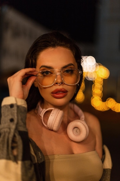 Modelo de moda mulher bonita com fones de ouvido rosa em cima e camisa usa óculos e caminha na cidade à noite com luz bokeh