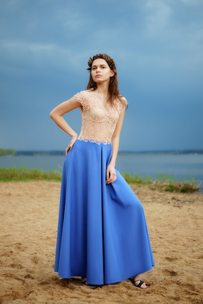 Foto modelo de moda solitário calmo andando na areia em um dia nublado em saia longa azul e blusa de renda.