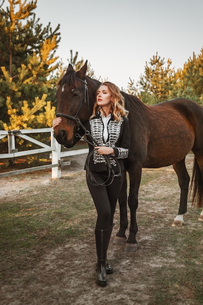 Foto modelo de mulher bonita de terno preto fica com um cavalo perto do estábulo