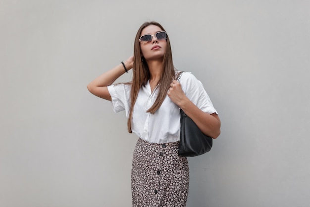 Modelo de mulher bonita de verão na moda com óculos de sol vintage em roupas da moda com uma camisa branca e uma saia com uma bolsa preta na moda fica perto de um prédio cinza na cidade
