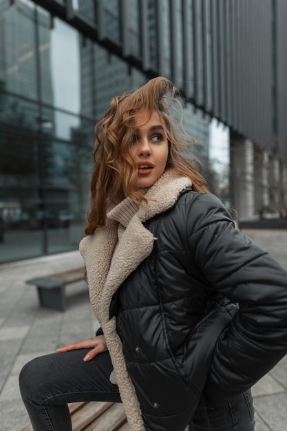 Modelo de mulher bonita elegante em roupas da moda de inverno com uma jaqueta e um suéter de malha com jeans posando na cidade perto do prédio de escritórios