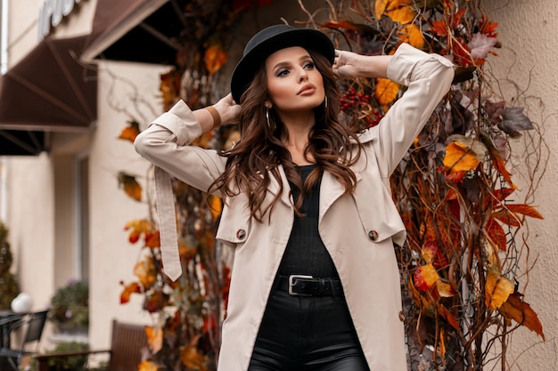 Modelo de mulher bonita elegante em roupas da moda elegantes com chapéu e casaco cinza andando na cidade com folhagem amarela de outono