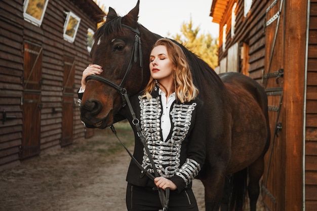 Modelo de mulher bonita em jaqueta preta está de pé com um cavalo perto do estábulo