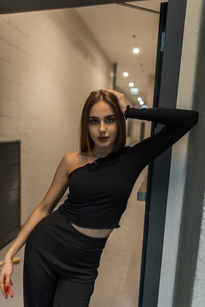 Modelo de mulher bonita na moda em elegantes roupas pretas elegantes está de pé perto da porta do prédio de escritórios