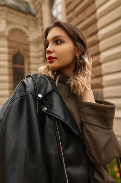 Modelo de mulher jovem e bonita com lábios vermelhos na moda jaqueta de couro preta e suéter verde caminha na cidade em um dia de outono