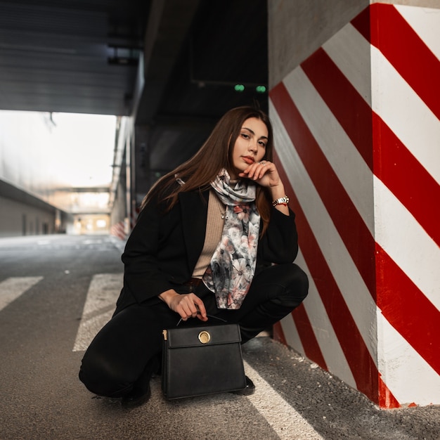 Modelo de mulher jovem e bonita elegante com roupas da moda com lenço de seda da moda com bolsa da moda de couro preto se passando perto de um pilar listrado no estacionamento da cidade. Linda garota descansa na rua. Senhora da beleza