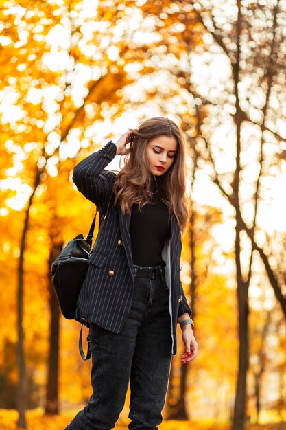 Foto modelo de mulher jovem e bonita elegante em um terno preto com um blazer, suéter e mochila da moda caminha em um parque de outono com folhagem de outono amarela ao pôr do sol