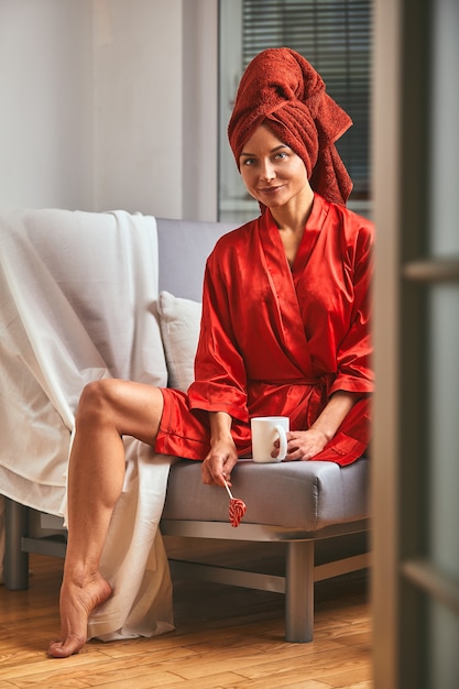 Modelo de roupão vermelho e toalha vermelha no sofá com uma caneca de café e um pirulito. O conceito de moda, o jogo de cores, o minimalismo. Descansar do trabalho, tempo de descanso.