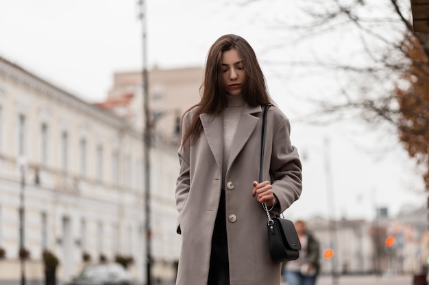 Modelo elegante con cabello largo castaño con un elegante abrigo posando en la ciudad en un día de otoño