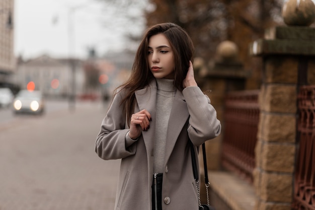 Modelo elegante con cabello largo castaño con un elegante abrigo posando en la ciudad en un día de otoño