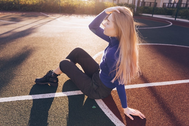 Modelo elegante de menina de cabelos compridos e suéter azul sentada em um parquinho sob os raios do sol da manhã
