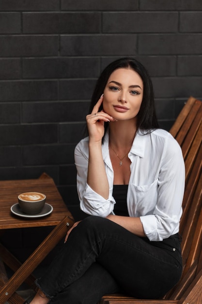 Modelo elegante e elegante em roupas casuais da moda com jeans e camisa branca, senta-se e descansa em um café na rua e bebe cappuccino perto de uma parede de tijolos pretos