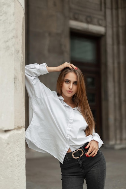 Modelo elegante e linda jovem em roupas casuais da moda com uma camisa branca e jeans preto fica perto de um prédio vintage na cidade Menina bonita