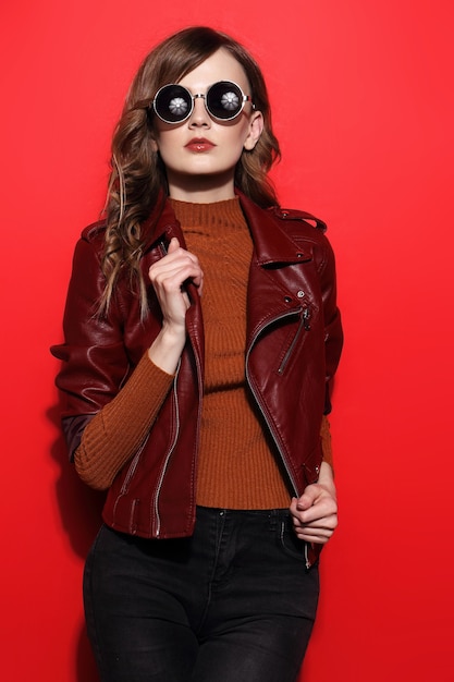 Modelo em óculos de sol, mulher jovem e bonita. jaqueta de couro, foto de estúdio, fundo vermelho