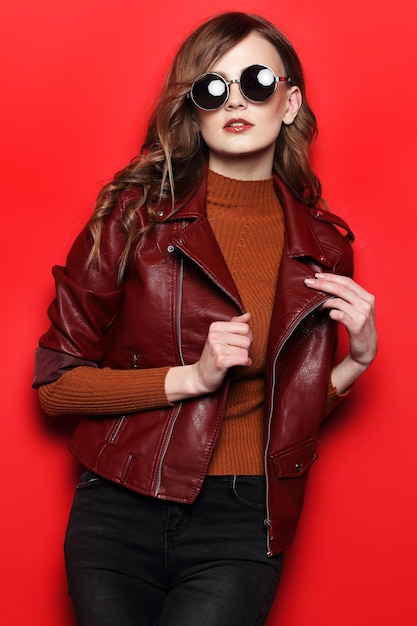Modelo em óculos de sol, mulher jovem e bonita. jaqueta de couro, foto de estúdio, fundo vermelho