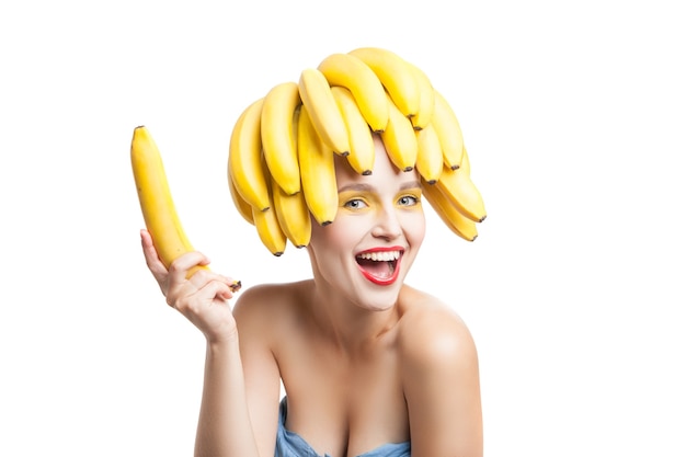 Modelo feliz com maquiagem e cacho de bananas na cabeça segurando na mão e olhando para a câmera.