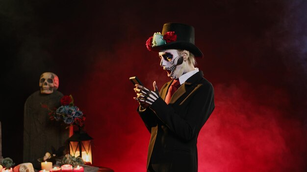 Modelo femenina de terror navegando por un sitio web de Internet en un teléfono inteligente, usando traje y sombrero para celebrar el día de los muertos. Dama de la muerte navegando en línea en teléfonos móviles, redes sociales. Disparo manual.