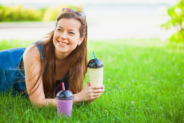 Modelo femenino atractivo se encuentran en el campo de hierba y beben batidos frescos. Mujer joven feliz con delicioso batido al aire libre.