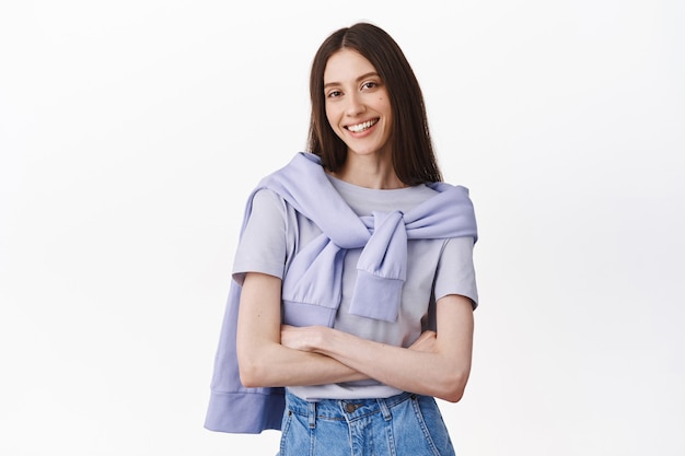 Modelo femenino confiado sonriente con los brazos cruzados en el pecho, pose de poder, luciendo asertiva y determinada al frente, de pie contra la pared blanca