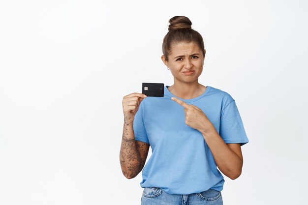 Modelo femenino escéptico señalando con el dedo a la tarjeta de crédito, frunciendo el ceño y haciendo muecas por disgusto, fondo blanco