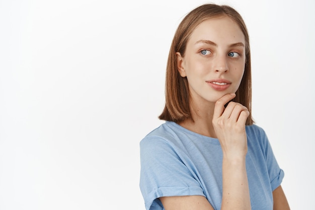 Modelo femenino joven atractivo con condición natural perfecta de la piel, rostro de mujer sin maquillaje y acné, sonriendo y mirando a un lado al anuncio de espacio vacío, pared blanca.