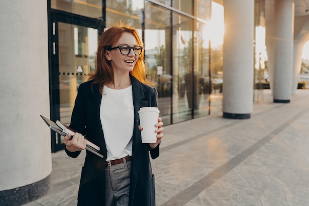 Modelo femenino sostiene el portátil con panel táctil y una taza de café para llevar plantea al aire libre durante el día soleado