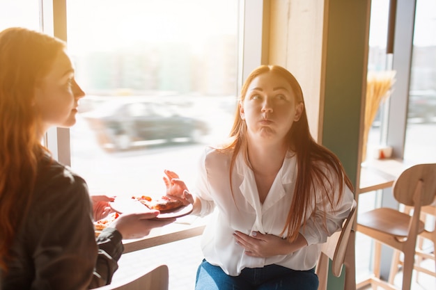 Modelo femenino toca el estómago y siente dolor. Joven comió mucha pizza en un café con su amiga.