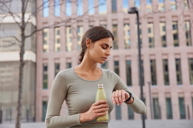 modelo feminina verifica o tempo no smartwatch bebe água doce fica com sede após o treinamento físico vestida em poses de roupas esportivas em um prédio antigo desfocado