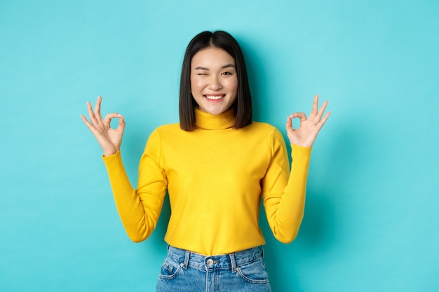 Modelo feminino asiático alegre mostrando gestos bem, sorrindo e parecendo impressionado, produto de elogio, em pé sobre um fundo azul.