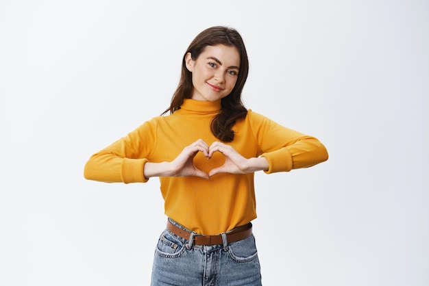Modelo feminino bonito sorrindo mostrando o gesto do coração da mão sobre o peito e olhando para a câmera fazendo confissão de amor em pé contra o fundo branco