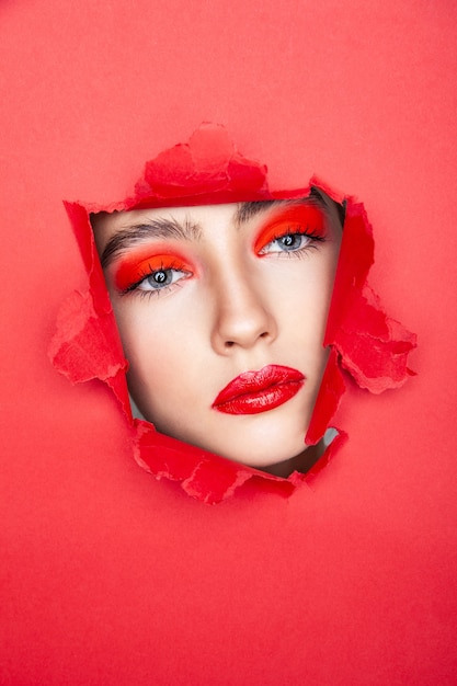 Modelo feminino encantador com maquiagem vermelha olhando para longe através de papel rasgado em estúdio