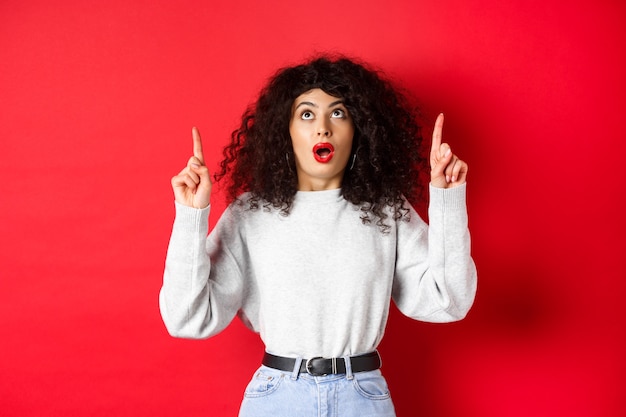 Modelo feminino europeu impressionado em roupas casuais, conferindo a promoção com o queixo caído, olhando e apontando os dedos para cima, mostrando o logotipo, em pé sobre fundo vermelho