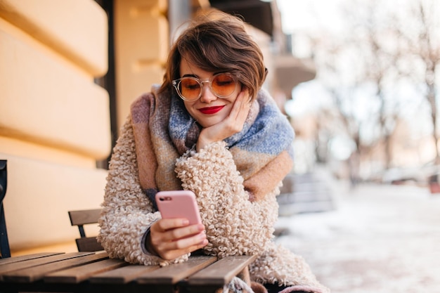 Modelo feminino jocund usando smartphone na rua Garota de cabelos curtos com casaco sentado no fundo da cidade