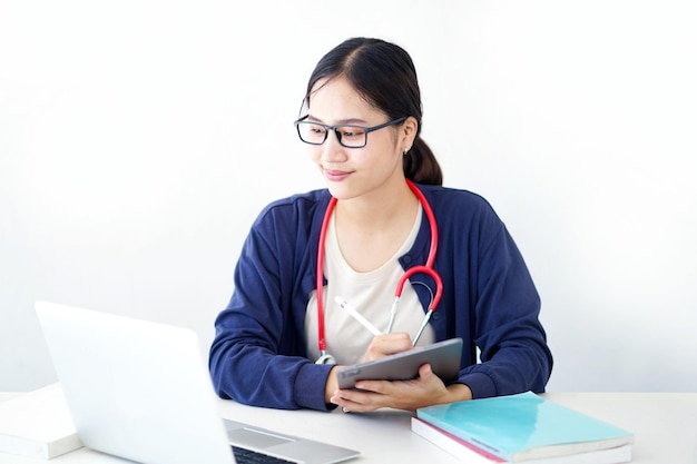 Modelo feminino jovem asiático atuando como médico olhando para laptop segurando um tablet