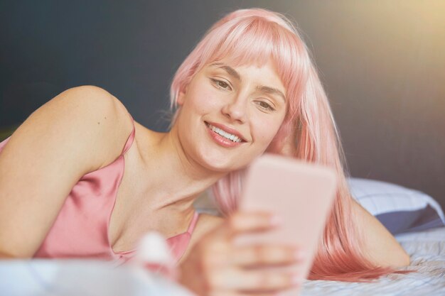 Modelo feminino sorridente com cabelo rosa lê mensagem no celular, deitado em uma cama confortável