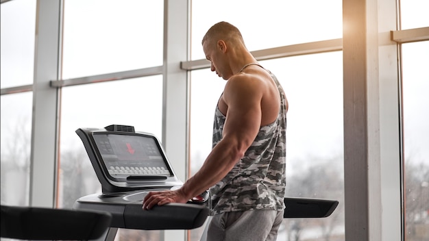 Modelo de fitness culturista atlético muscular corriendo gimnasio caminadora cerca de una ventana grande