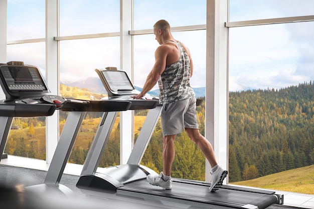 Modelo de fitness culturista atlético muscular corriendo gimnasio caminadora cerca de una ventana grande
